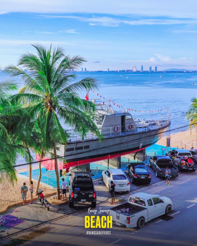 The image is of a military ship monument ship docked at Bang Saray Beach Road. The scene includes palm trees, the sea, and a cloudy sky. 