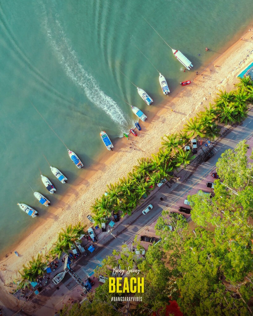 The image shows Bang Saray Beach, lined with fishing and tourist boats along the shore. It presents a picturesque aerial perspective of the sandy beach exuding a calm and soothing ambiance.