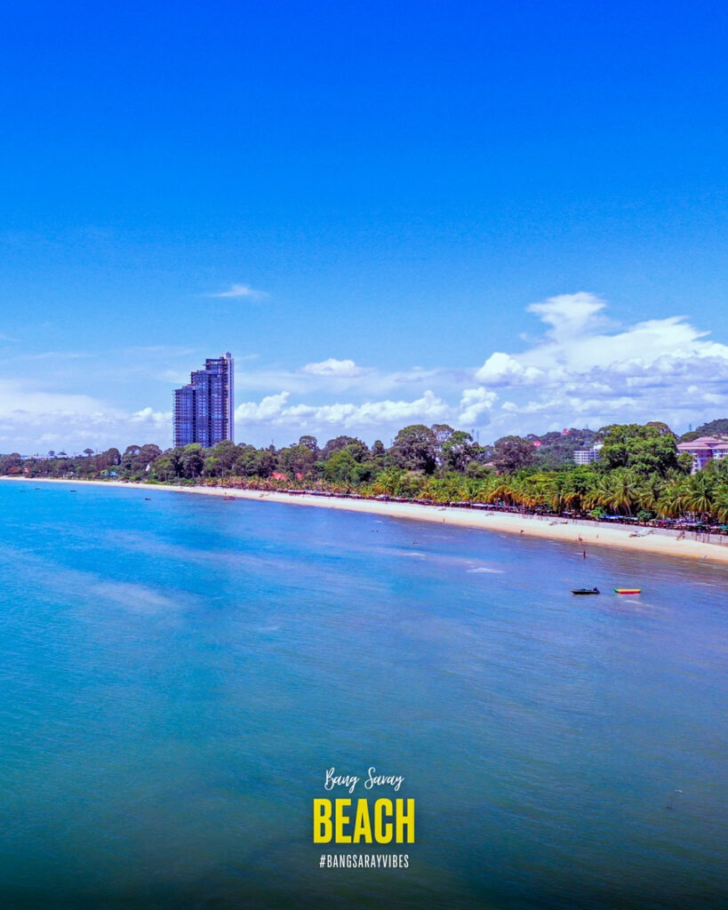 The drone picture showcases Bang Saray Beach and the Del Mare condominium, capturing the outdoor landscape featuring the sky, water, trees, and coastline. It presents a tropical beach environment blending nature with urban development.