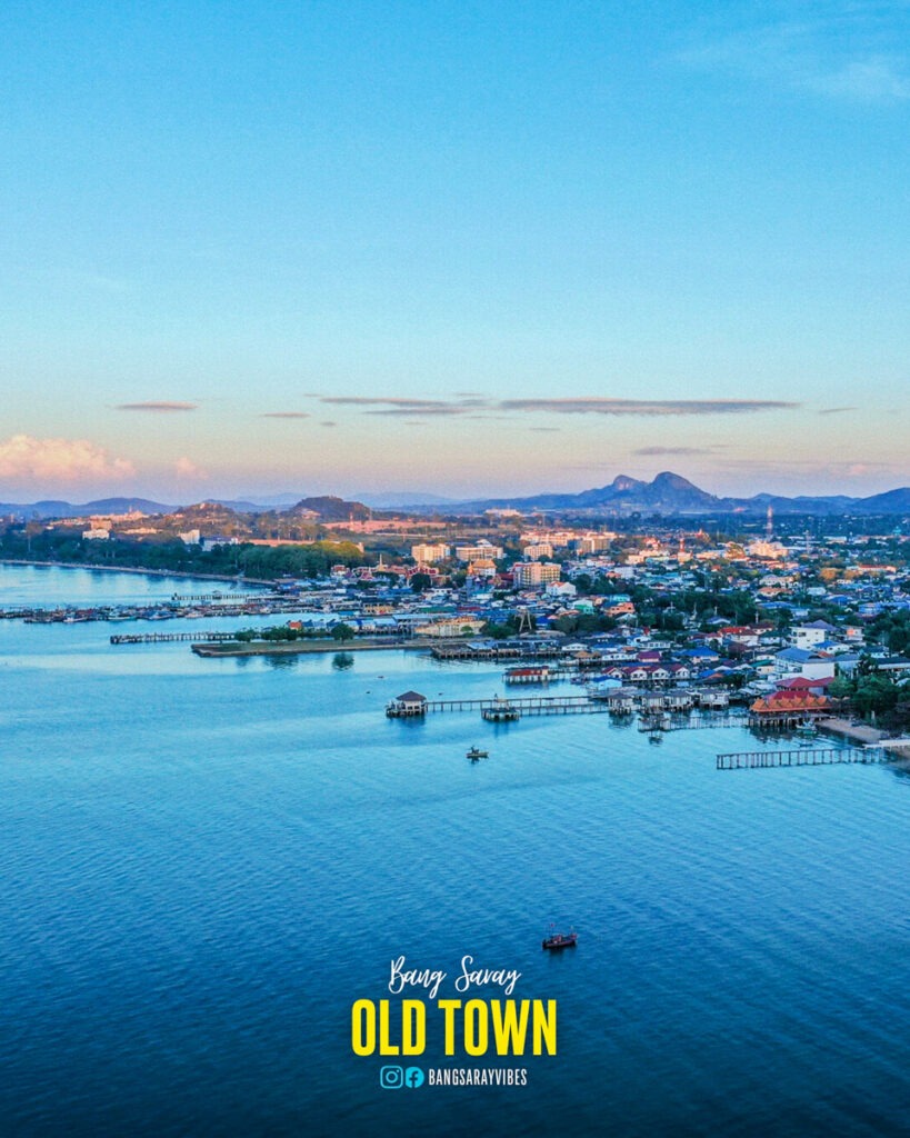 Aerial view of Bang Saray Old Town along the coastline with Thai traditional buildings, piers, and fishing boats on calm blue waters under a clear sky.