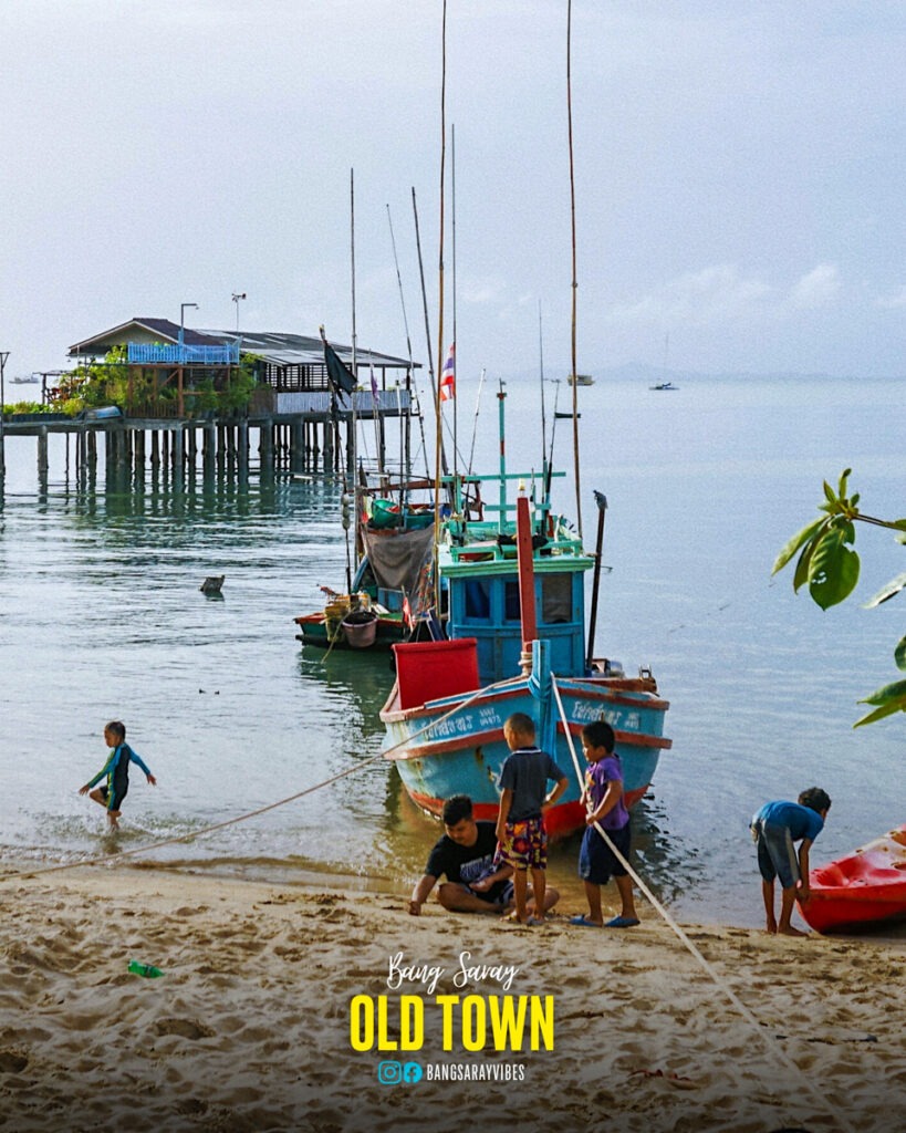 The image shows kids playing near a traditional Thai fishing boat on the beach in Bang Saray Old Town, located in the Chonburi province of Thailand.